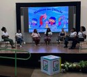 Para cego ver: Foto mostra mulheres sentadas em semi círculo em um palco; ao fundo, painel luminoso escrito meninas na ciência 