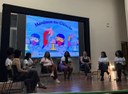 Para cego ver: Foto mostra mulheres sentadas em semi círculo em um palco por outro ângulo; ao fundo, painel luminoso escrito meninas na ciência