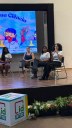 Para cego ver: Foto mostra mulheres sentadas em semi círculo em um palco por outro ângulo; ao fundo, painel luminoso escrito meninas na ciência