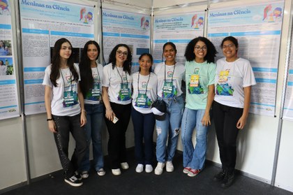 Para cego ver: Foto mostra grupo de mulheres e meninas posicionadas lado a lado. Elas usam camiseta e crachá de evento. Ao fundo, diversos posters de apresentação de pesquisas