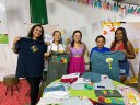 Para cego ver: cinco mulheres compõem a foto. Elas mostram camisetas produzidas pelo projeto. À frente delas, uma mesa com diversos produtos feitos por elas