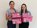 Para cego ver: Foto mostra duas mulheres centralizadas na imagem. Elas seguram caixas cor de rosa com o nome "fluxo sem tabu", onde serão disponibilizados os absorventes 